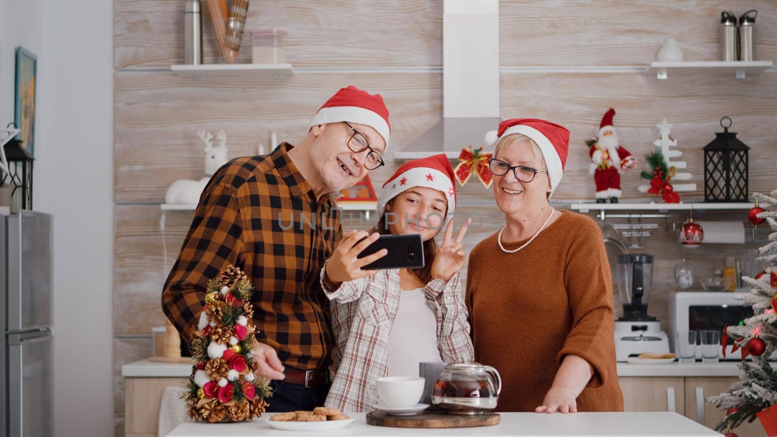 Happy grandparents standing at table in xmas decorated kitchen taking selfie using smartpgone during winter season. Family wearing santa hat making funny expressions enjoying christmas holiday
