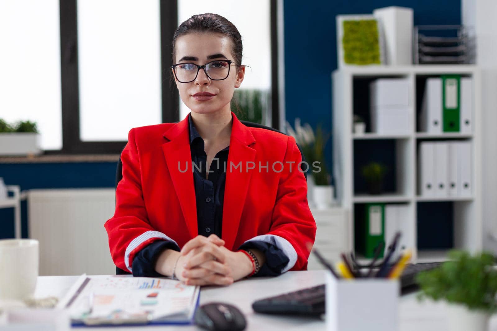 Portraif of confident businesswoman sitting at desk in corporate office workplace building, with finger crossed wearing glasses looking at camera. Financial charts and graphs.