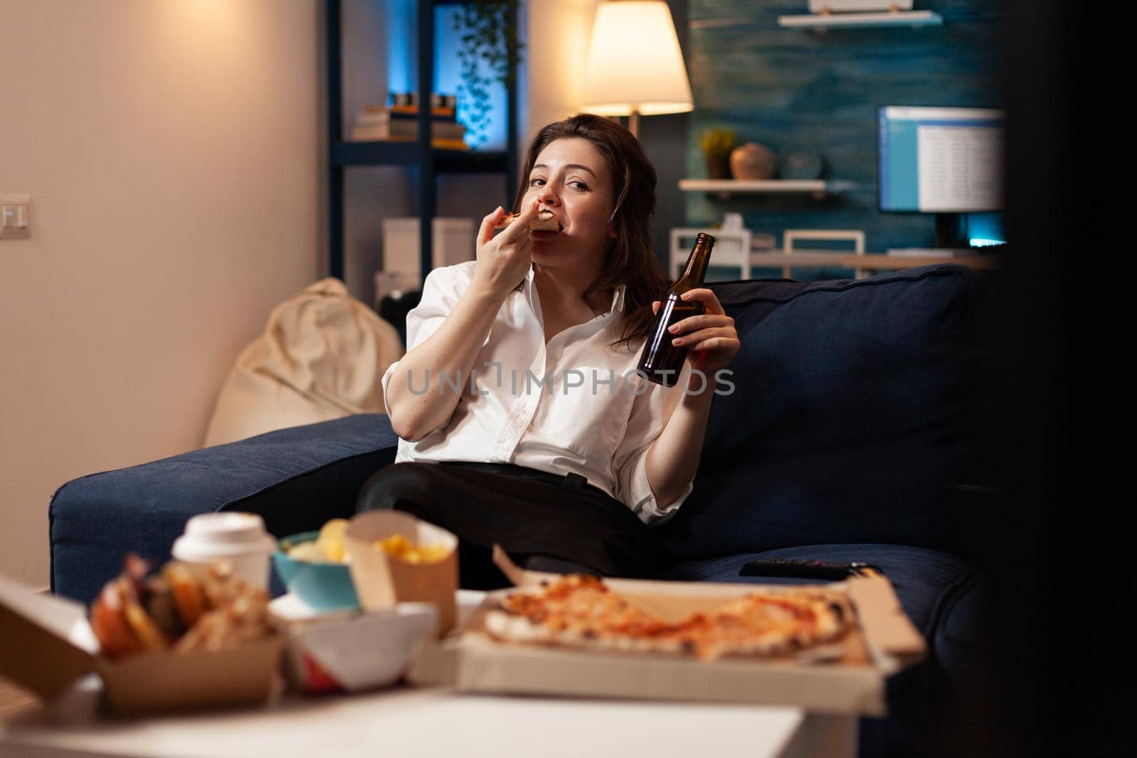 Happy woman eating tasty delicious delivery pizza slice relaxing on sofa watching comedy movie on television in living room. Caucasian female enjoying takeaway fast home delivered in evening