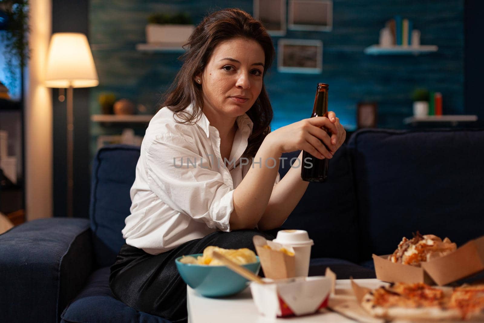 Portrait of cheerful woman sitting on couch smiling at camera while enjoying home-delivered late at night in living room. Caucasian female holding beer bottle. Takeaway food delivery
