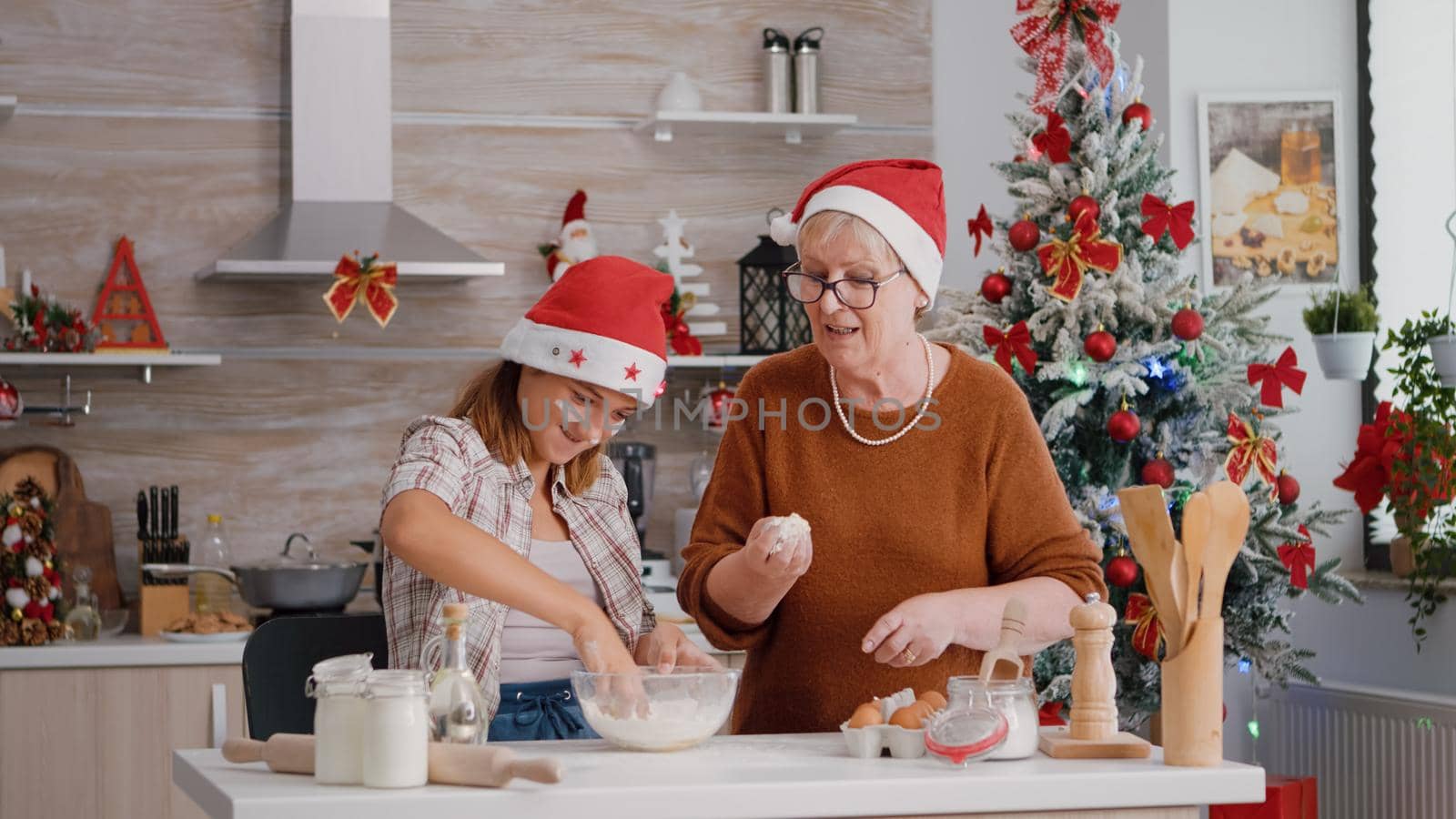 Grandparent helping grandchild preparing homemade traditional cookie dough in culinary kitchen during christmas holiday. Family wearing xmas hat enjoying cooking together making delicious dessert