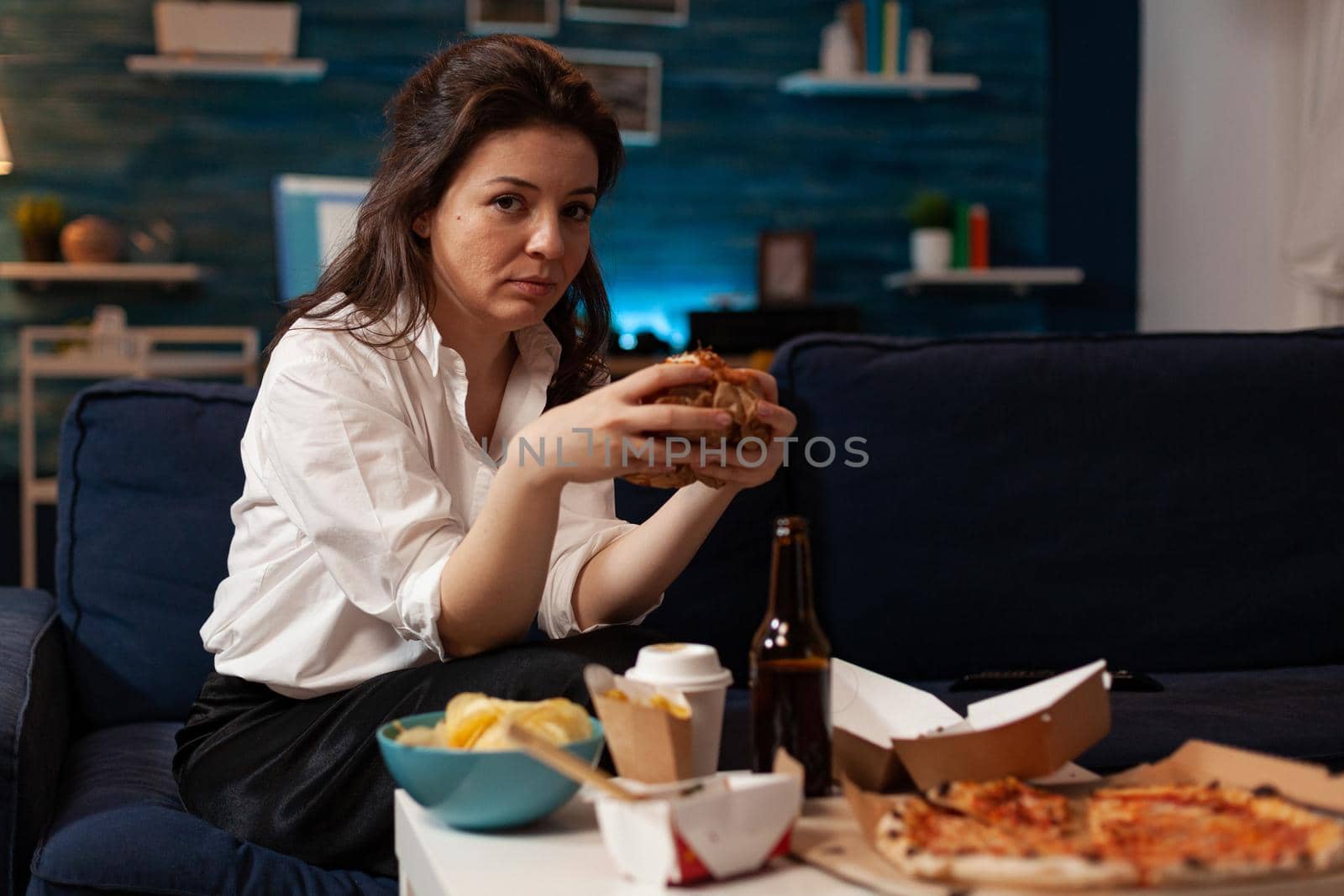 Portrait of relaxed woman looking into camera holding tasty burger in hands during fast food night in living room. Caucasian female enjoying delicious home delivered junk-food. Takeaway delivery