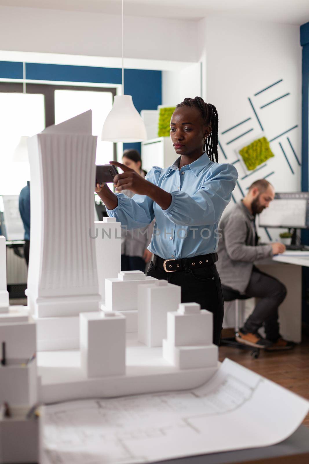 Woman of african american ethnicity working as architect with building plans maquette looking at smartphone for creative design. Worker viewing construction project for industry