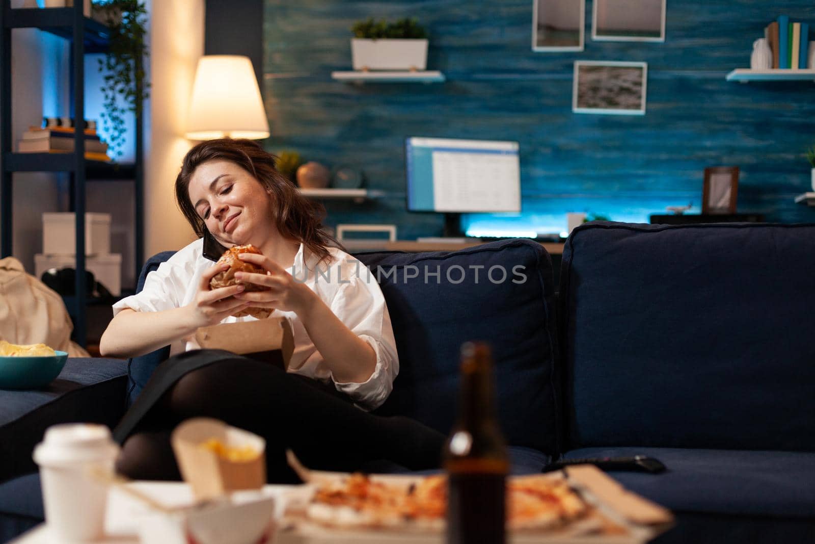 Woman talking at phone with friends while holding tasty delicious burger in hands relaxing on couch in living room late at night. Caucasian female enjoying tajeaway junk food home delivered