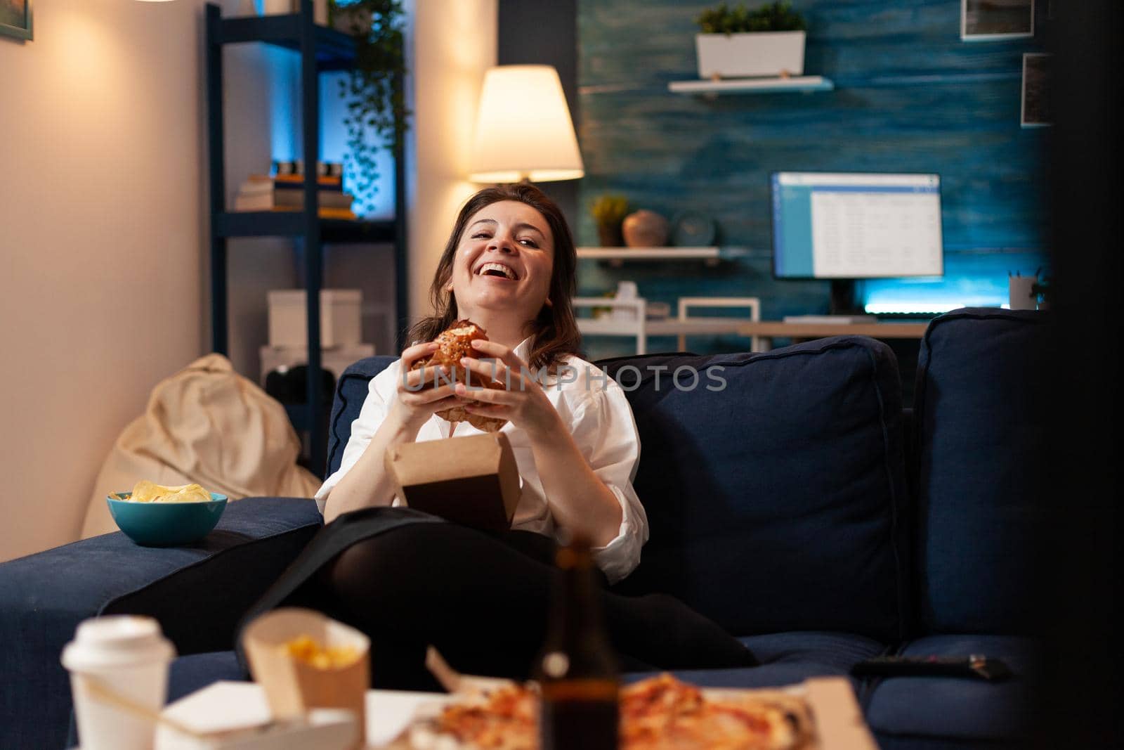 Caucasian female laughting while watching entertainment movie series on television eating tasty delicious fast food burger. Woman enjoying unhealthy takeaway food home delivered. Lunch meal order