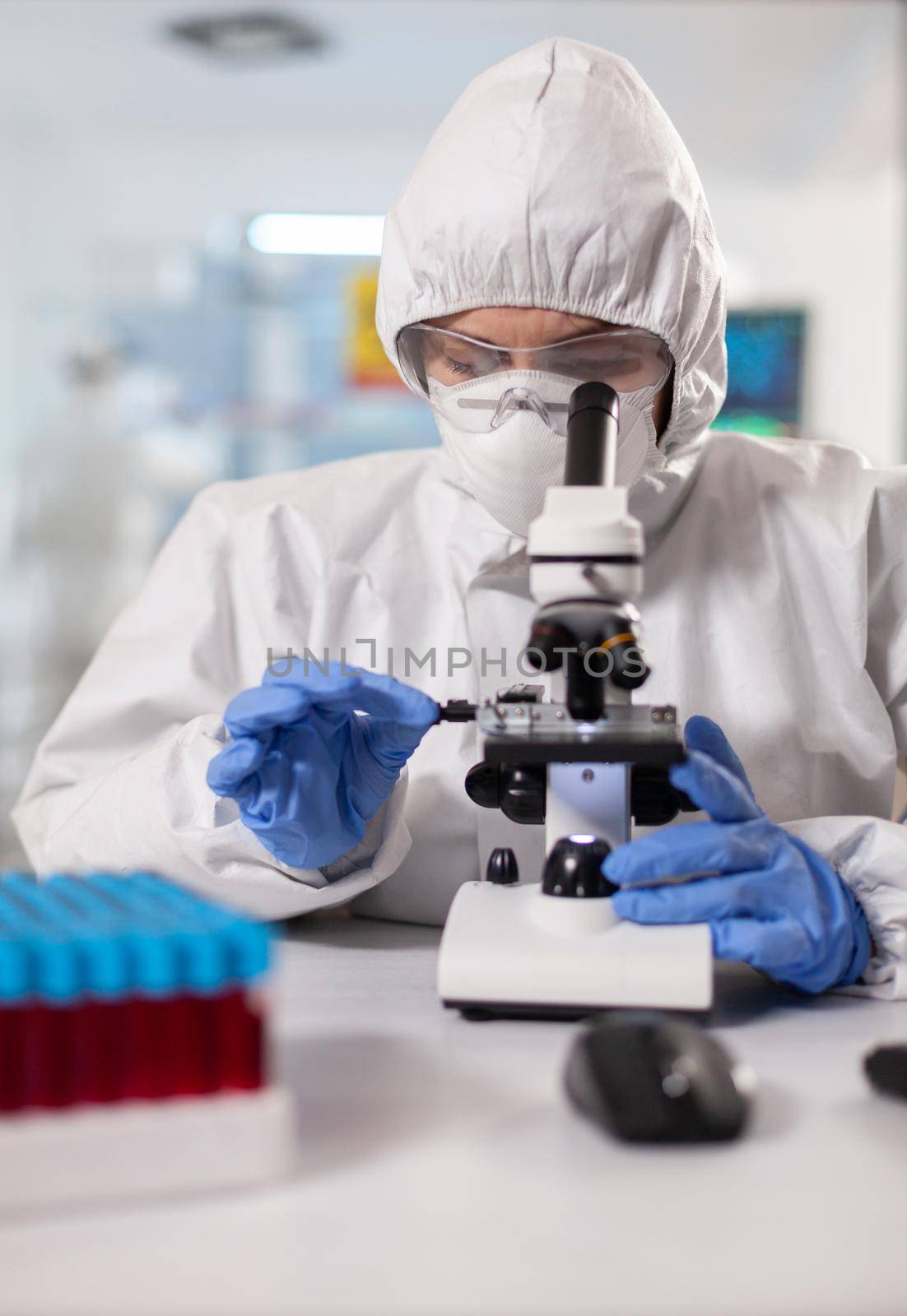 Genetic scientist looking at sample through microscope dressed in ppe suit. Chemist in coverall working with various bacteria, tissue blood samples for antibiotics research.