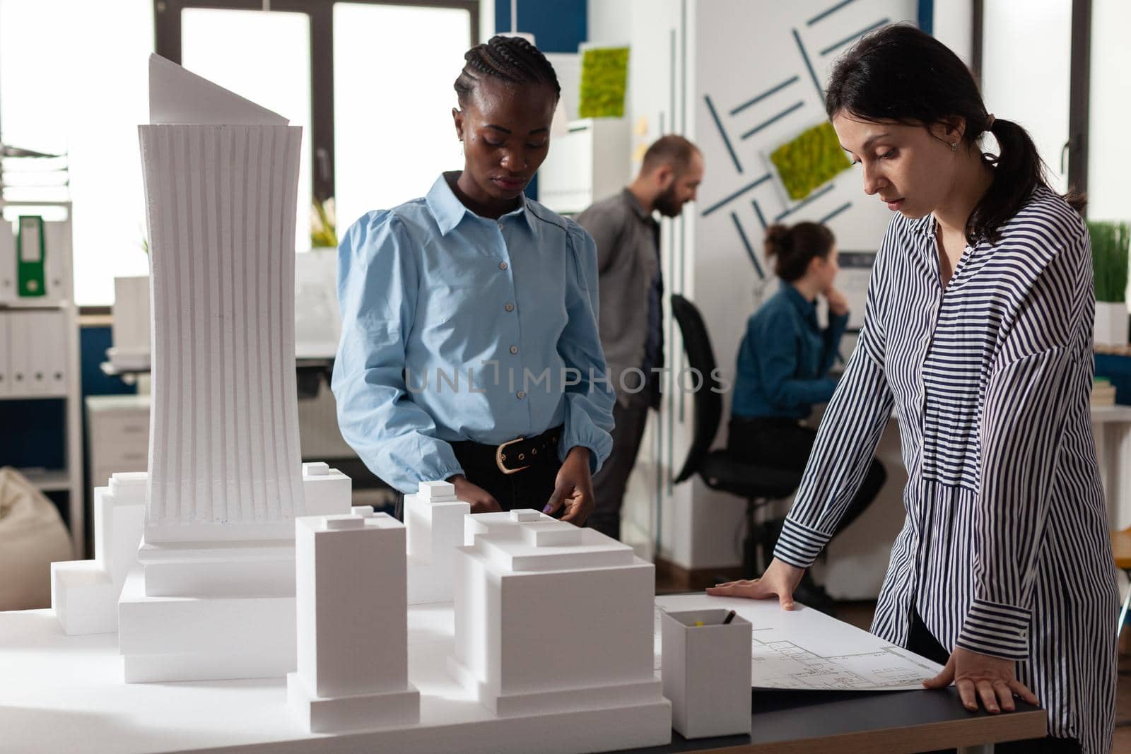 Multi ethnic women working as professional architects partners for designing blueprint plans. Colleagues standing at building model maquette urban industry design on desk.