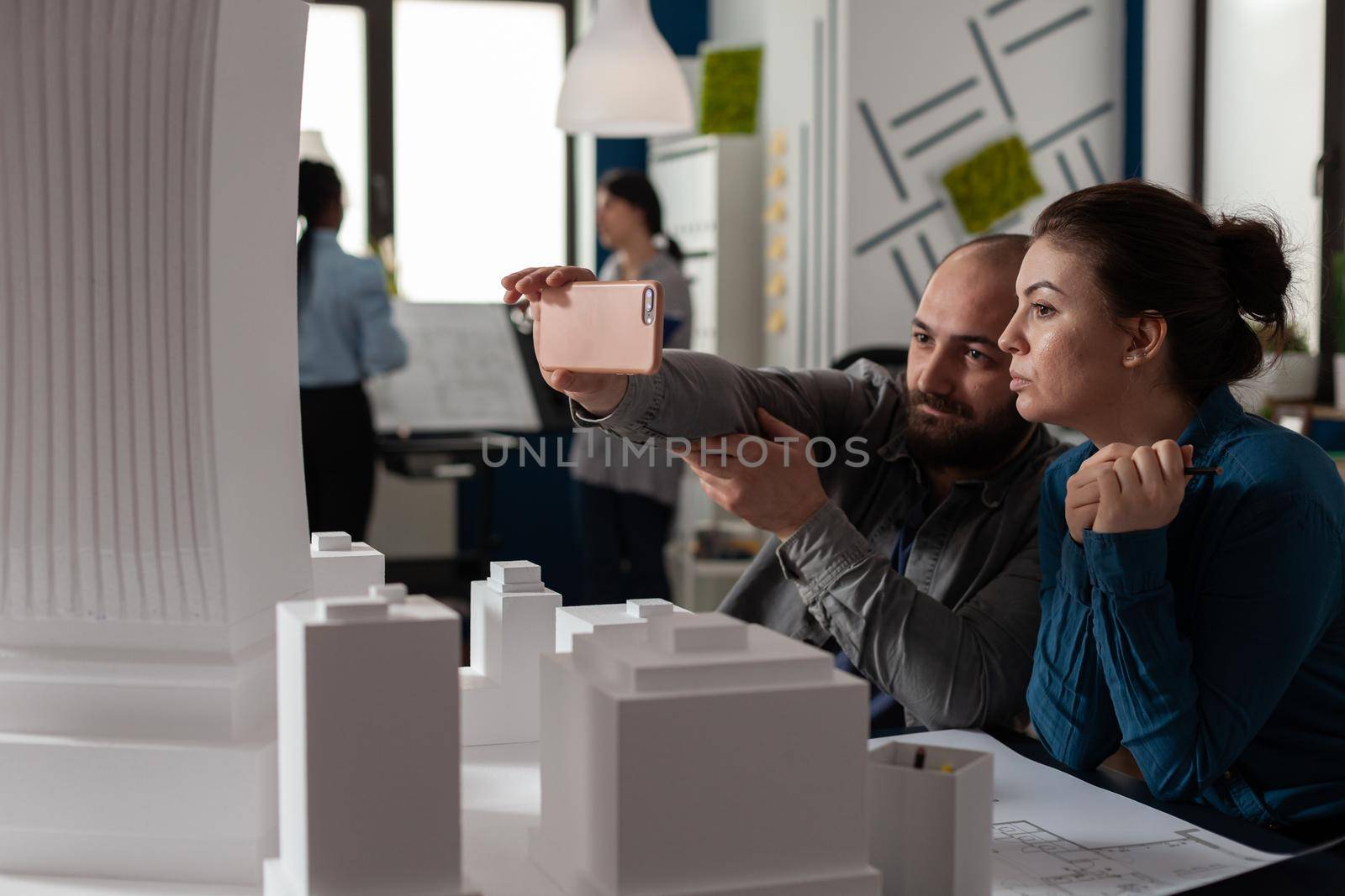 Professional architect colleagues work on smartphone sitting at desk while looking at building model maquette. Engineering workmates talking about construction industry project