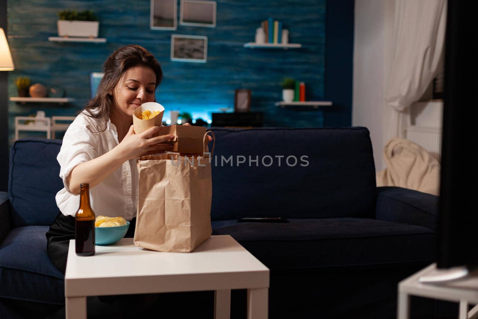 Smiling cheerful woman unpacking fast food meal delivery sitting on sofa during junk-food home-delivered in living room. Caucaisan female enjoying takeaway service lunch in evening