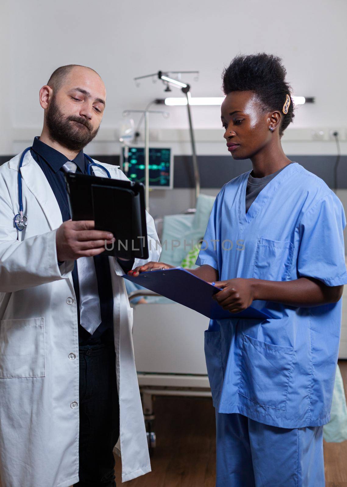 Physician doctor with asisstant with black skin checking illness recovery treatment using tablet computer during medical consultation in hospital ward. Hospitalized patient sick woman resting in bed