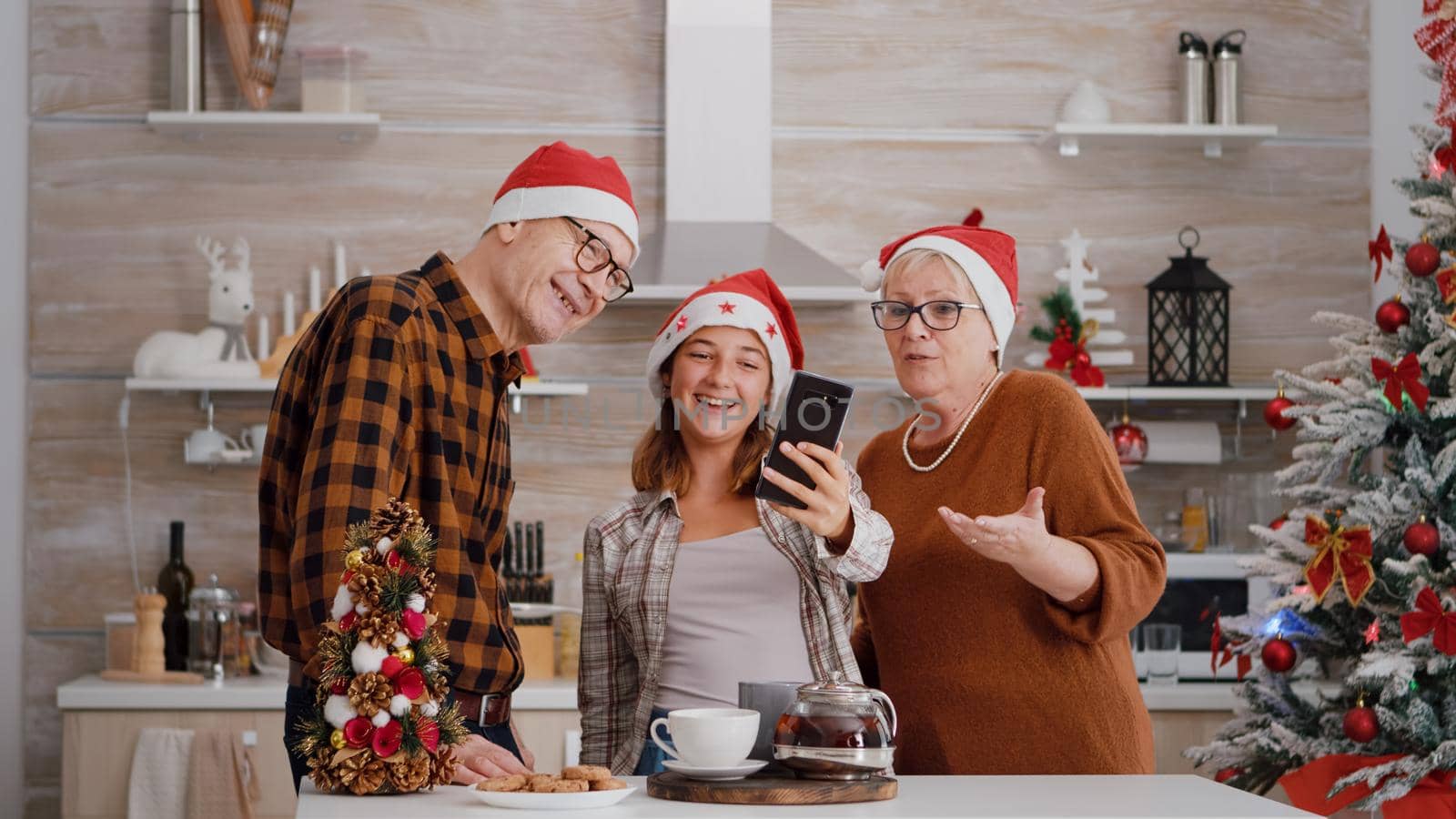 Grandparents with granddaughter greeting remote mother celebrating xmas holiday during online videocall meeting using mobile. Family wearing santa kat enjoying christmas winter season