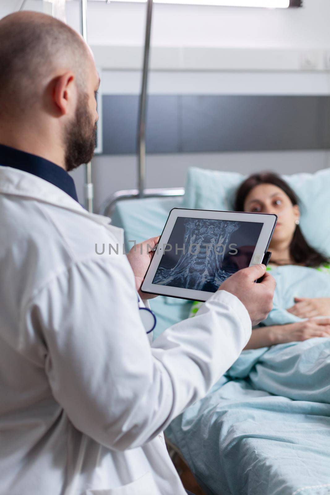 Specialist doctor monitoring sick woman explaining bones radiography using tablet computer working in hospital ward. Patient resting in bed recovering after sickness surgery
