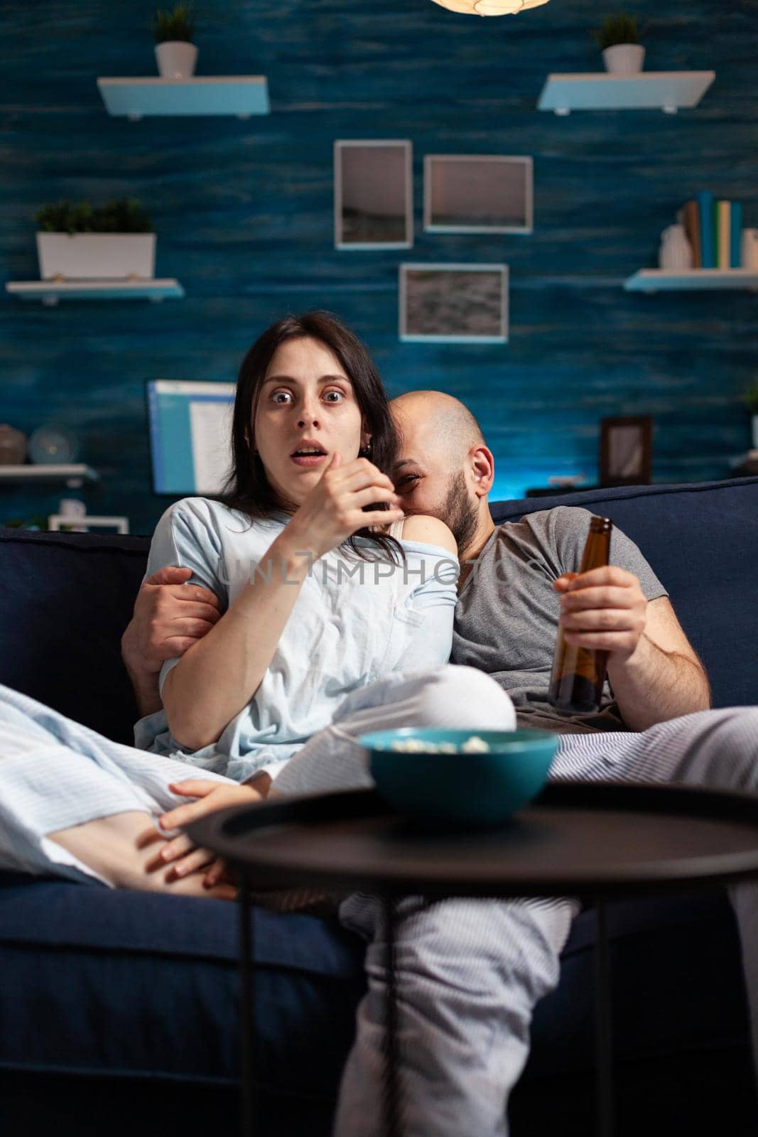 Confused shocked couple watching TV movie at night and eating popcorn, drinking beer having fear facial expression. Focused astonished people sitting on confortable couch chilling together