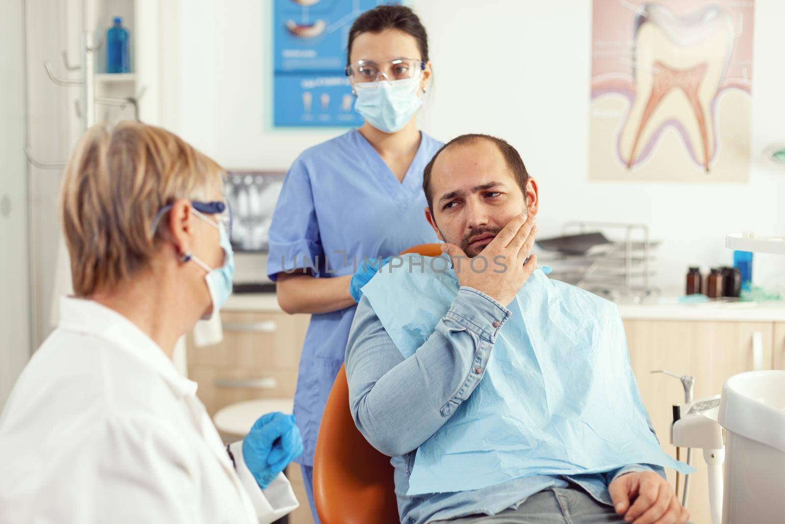Sick man having symptoms of tooth pain holding hand on cheek while talking with senior doctor. Patient explaining dental problem to stomatologist woman sitting in stomatology hospital clinic office