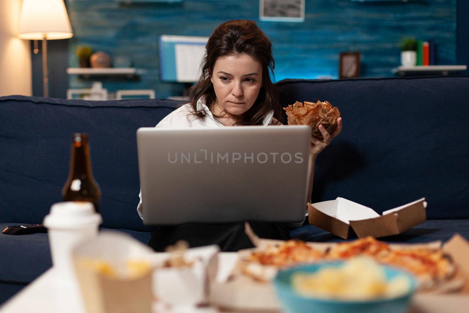 Caucasian female sitting on sofa eating tasty delicious burger while working on laptop computer in living room late at night. Woman enjoying takeaway food home delivered. Fastfood lunch meal order