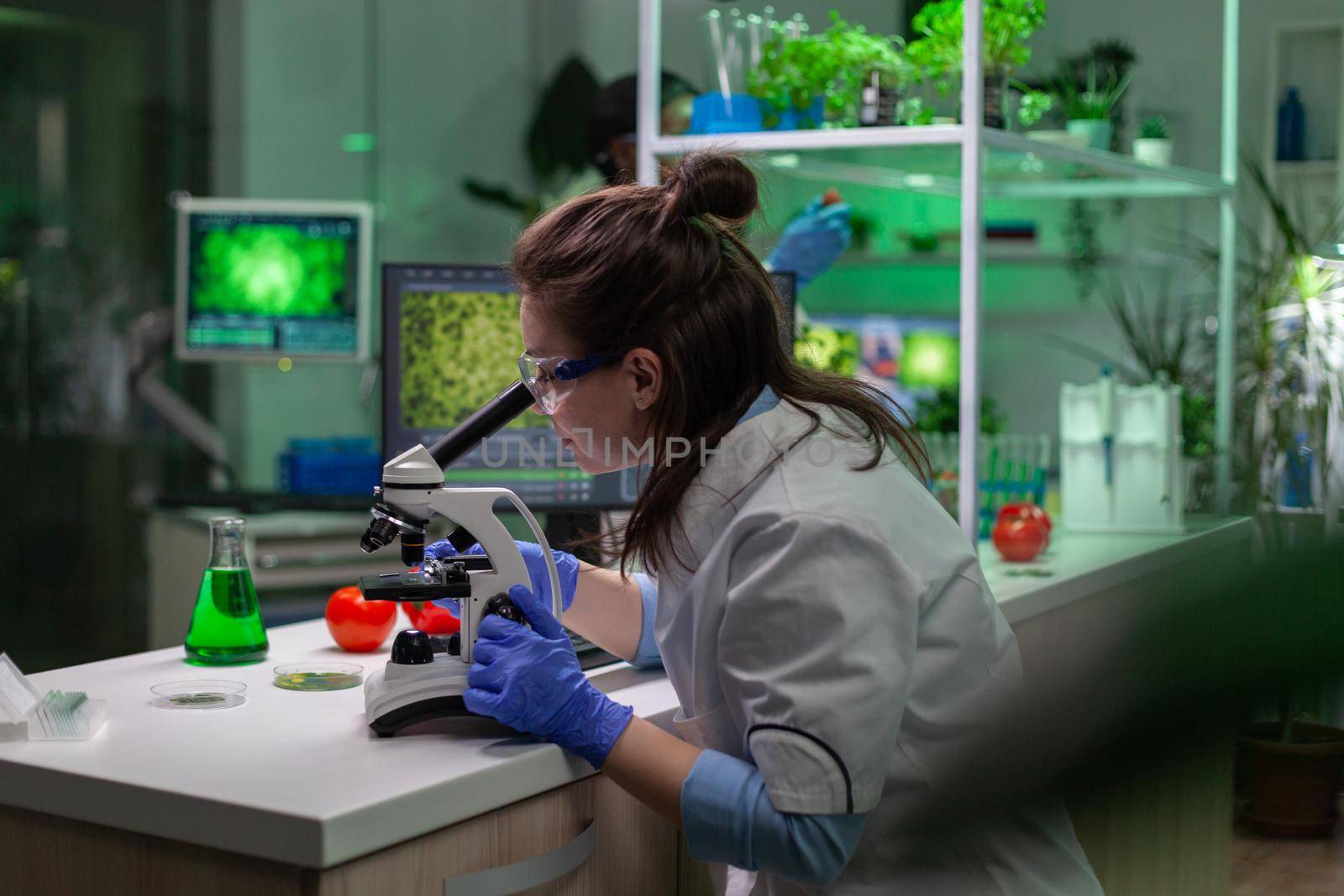 Biologist scientist in white coat working in expertise laboratory looking into microscope analyzing organic gmo leaf. Specialist researcher doing biochemistry experiment.