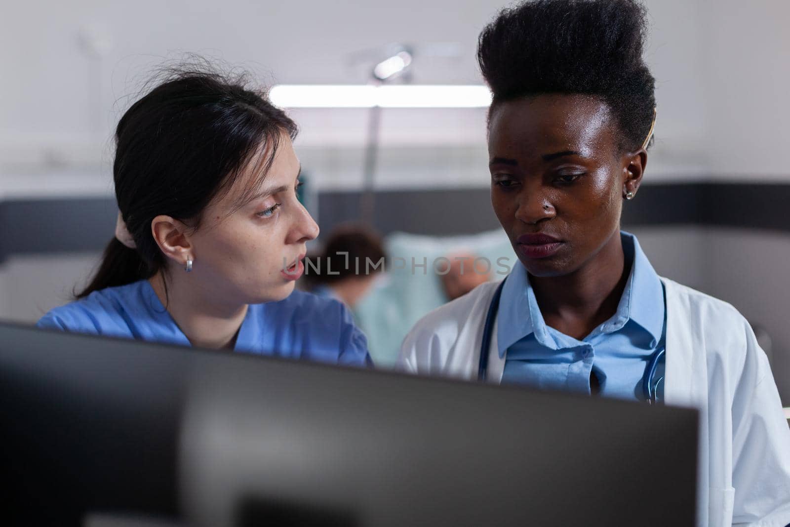 Medical team looking at computer analyzing healthcare treatment working in hospital ward. In background doctor discussing with sick man patient, recovering after illness surgery