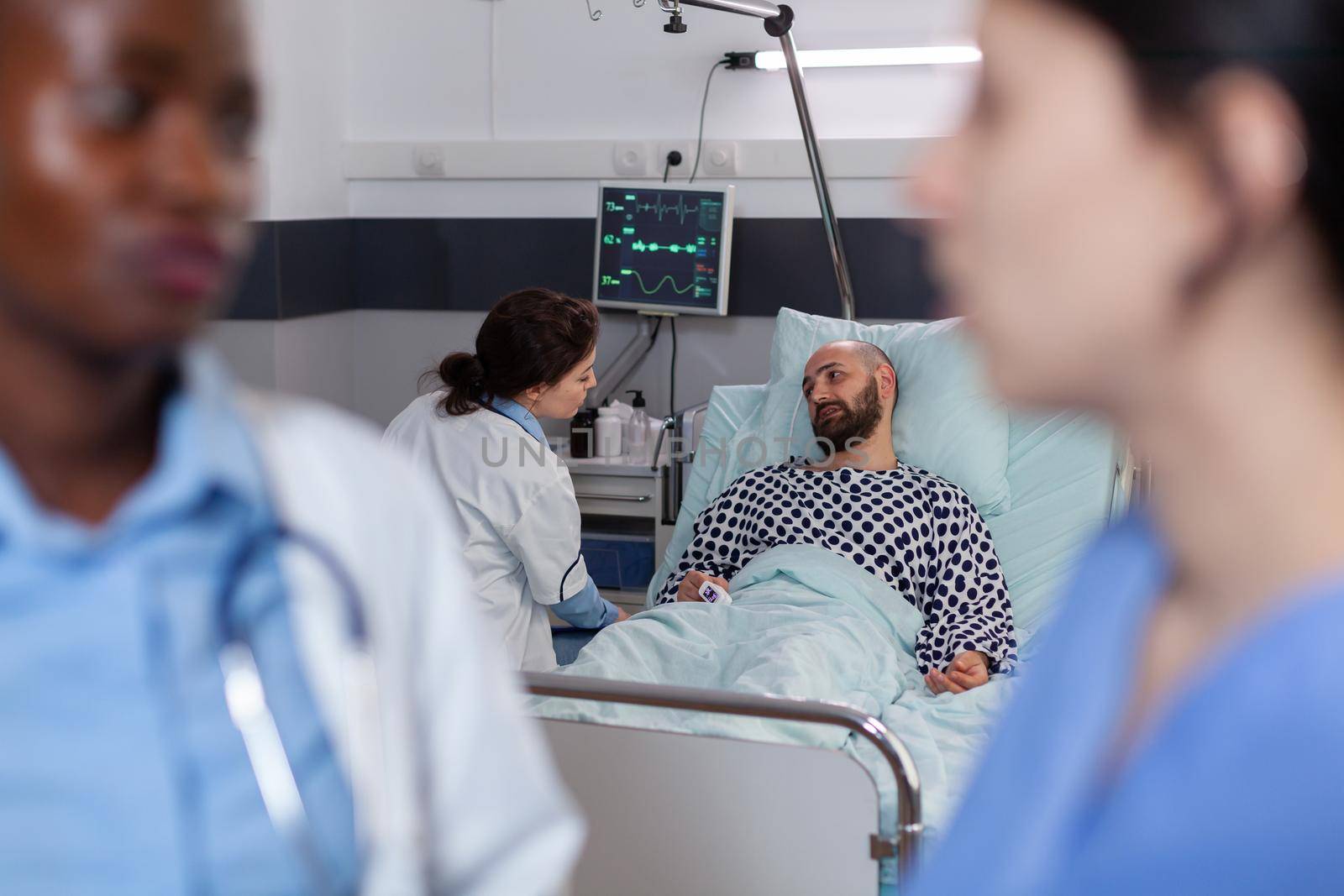 Physician doctor with black skin talking with medical asisstant during recovery consultation working in hospital ward. Sick man patient resting in bed explaining disease symptom to practitioner