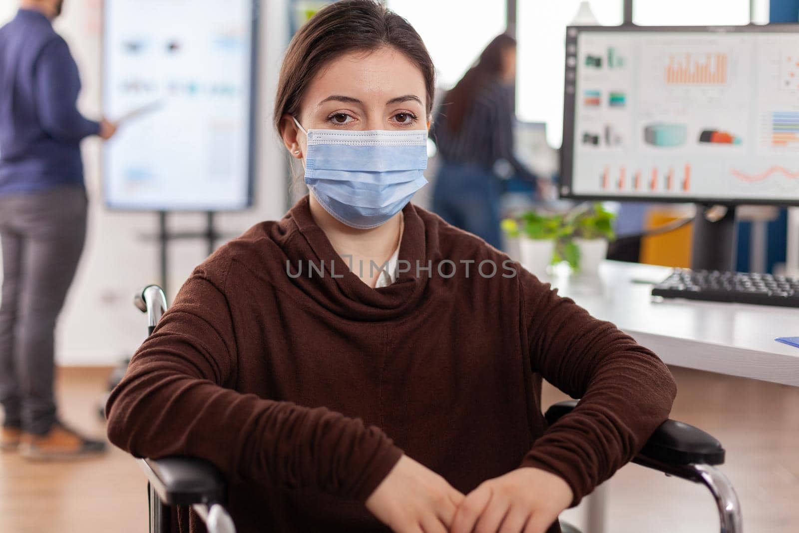 Woman employee with disabilities, invalid, handicapped paralized with protection mask against coronavirus looking at camera sitting immobilized in wheelchair in new normal business office room.