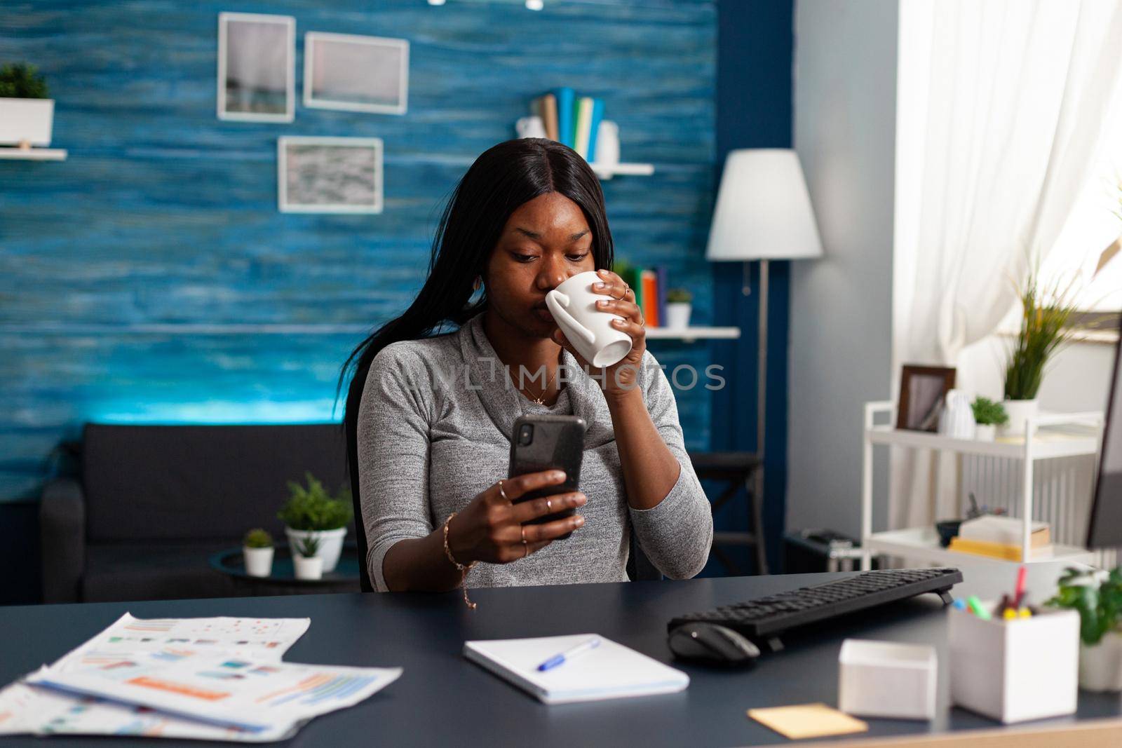 Black woman holding smartphone in hands chatting with people browsing communication information sitting at desk in living room. African american student looking on social media