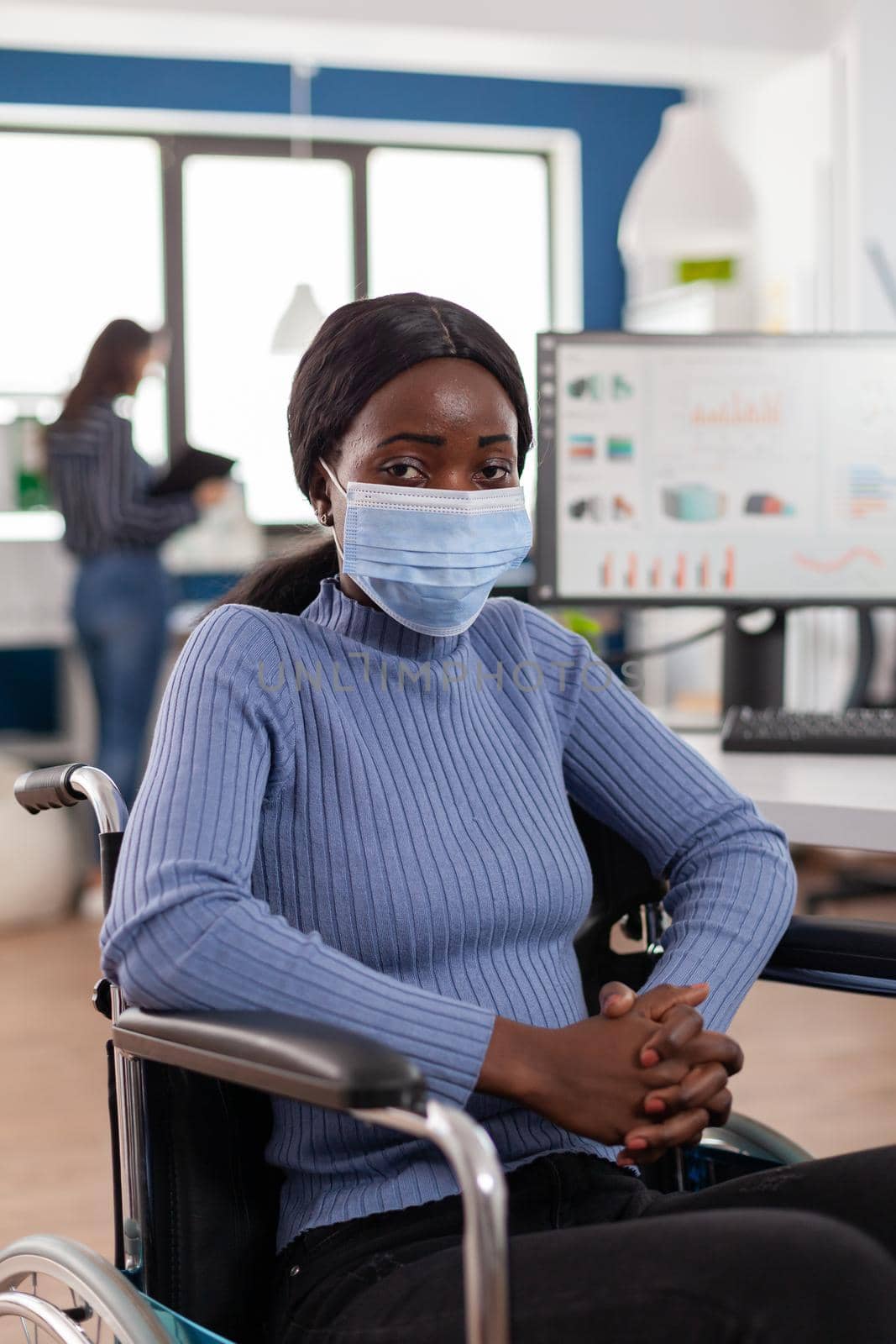 African invalid handicapped woman employee with protection mask sitting immobilized paralized in wheelchair looking lost, new normal business office during covid 19 global pandemic working with team.