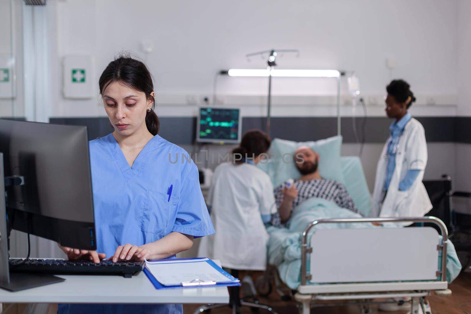 Woman asisstant typing medical recovery on computer while in background practitioner doctor monitoring sick man explaining disease symptom. Hospitalized patient having respiratory disorder