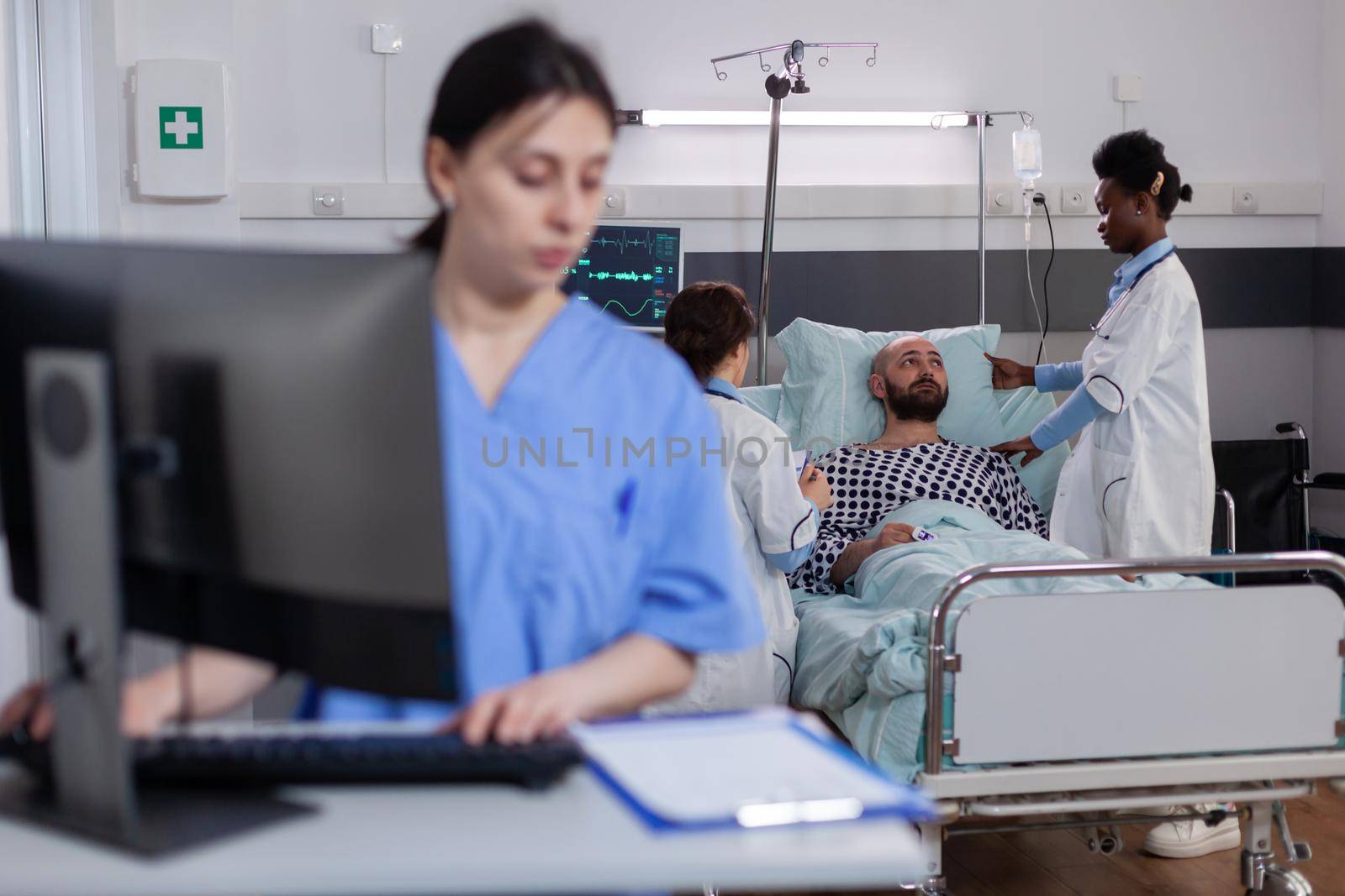 Front view of asisstant typing medical expertise on computer while in background black practitioner doctor discussing with sick man healthcare treatment. Hospitalized patient having respiratory disorder