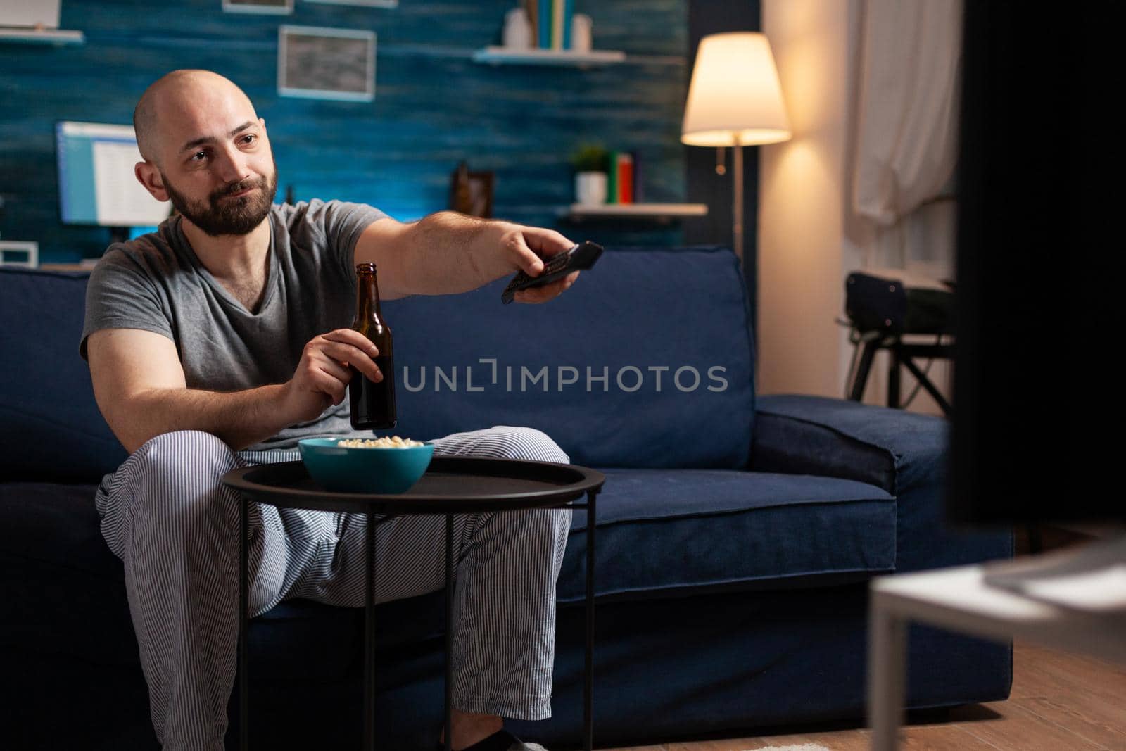 Excited relaxed man spending free time watching TV entertainment series eating popcorn and drinking beer. Funny amused enjoying evening at home sitting on comfortable couch dressed in pajamas