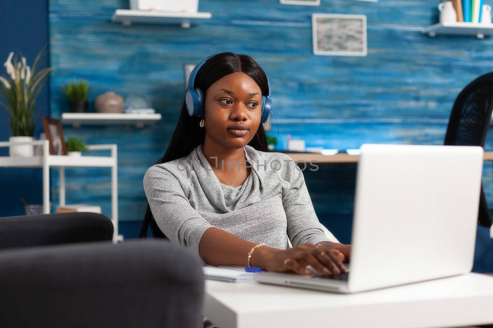 African american student wearing headphones during education webinar remote working at communication blog article. Woman with black skin browsing social media information on internet