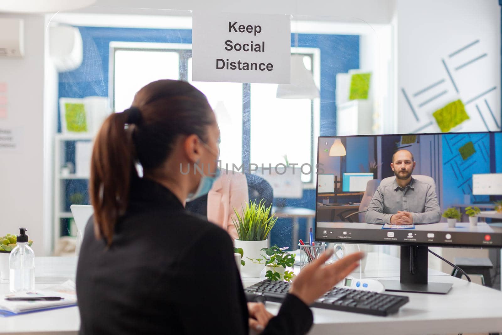 Back shot of businesswoman wearing medical face mask talking on videocall meeting with remotely team. Executive freelancer working in new normal company office during coronavirus pandemic.