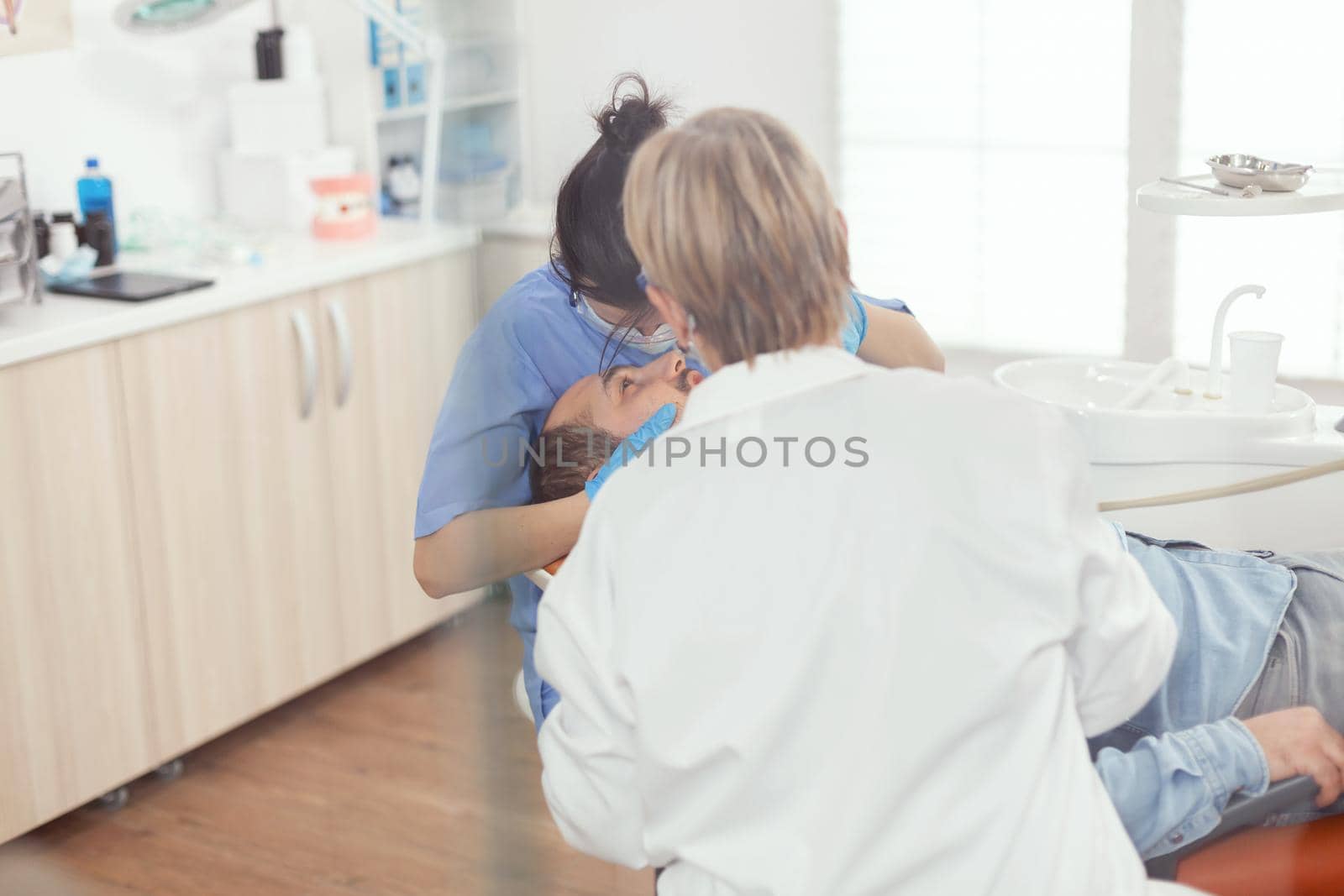 Sick patient sitting on dental chair with open mouth during stomatology procedure. Senior woman stomatologist and hospital nurse with masks holding sterilized tools checking teeth health