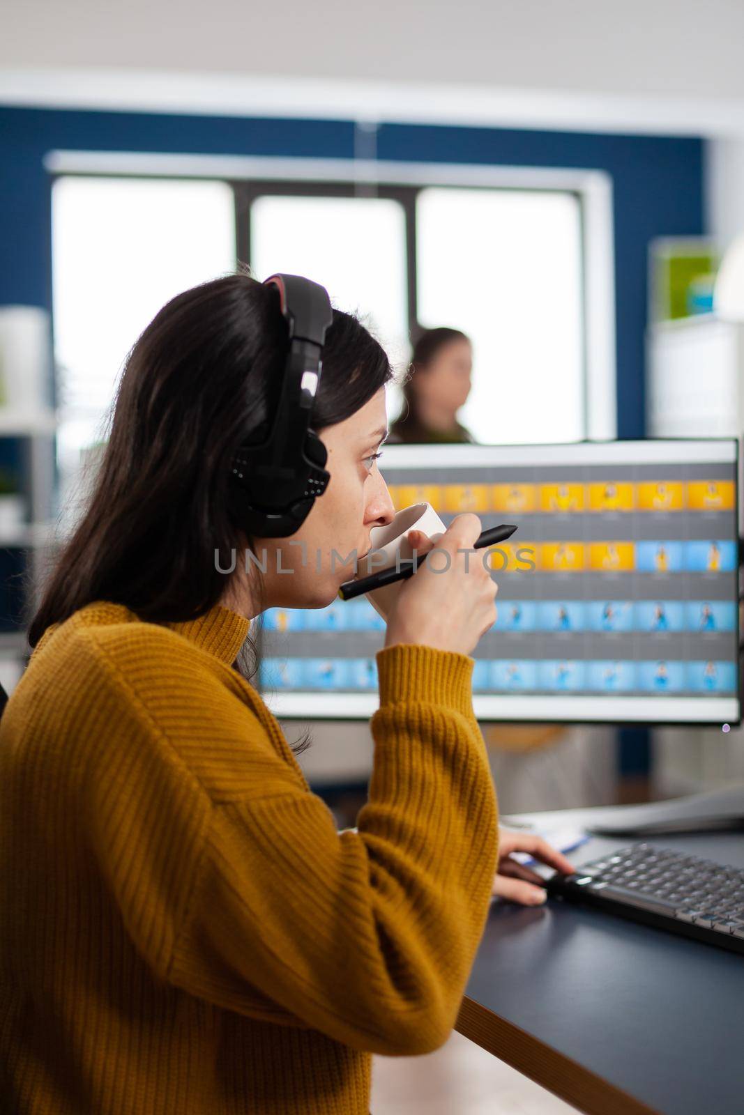 Photo editor with headphones retouching image using stylus pen, drinking coffee, working on PC with two displays. Woman retoucher editing in software app with graphic tablet sitting in creative agency