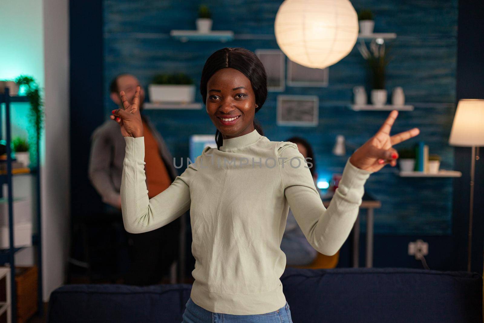 Potrait of afro american woman smiling into camera showing victory hand sign late at night in living room. In background multiethnic friends gathering together having fun during wekeend party.