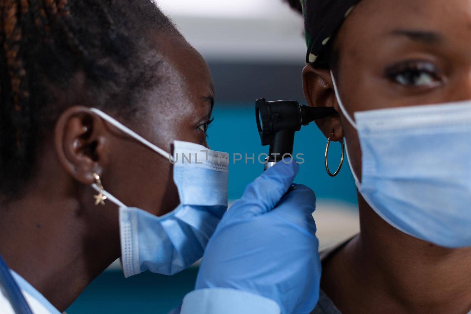 Closeup of african american otologist doctor with protective face mask against coronavirus checking patient ear using medical otoscope during clinical appointment in hospital office. Otoscopy tool