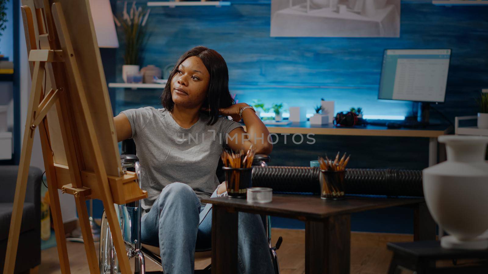 Paralyzed black artist sitting in wheelchair drawing on canvas with easel using pencil. African american woman with disability working on beautiful masterpiece of authentic white vase