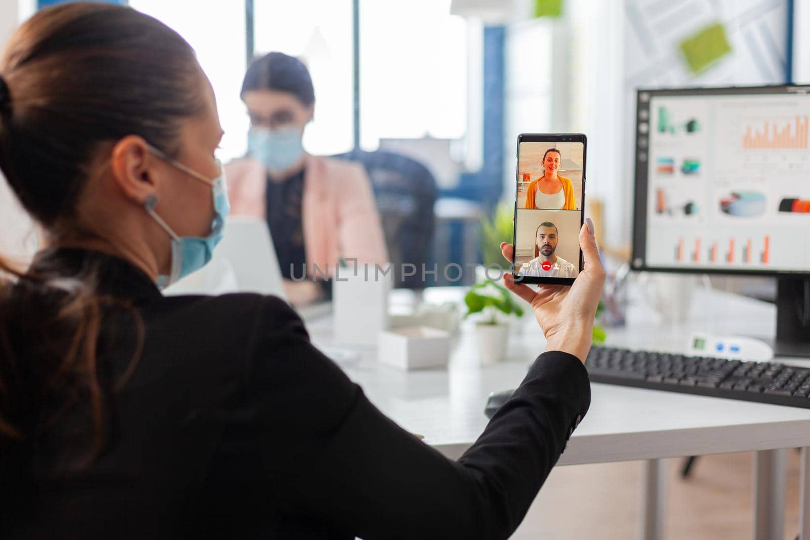 Back view of woman with face mask talking during business video call keeping social distancing at workplace, safety precaution against infection with covid19 flu at workplace.