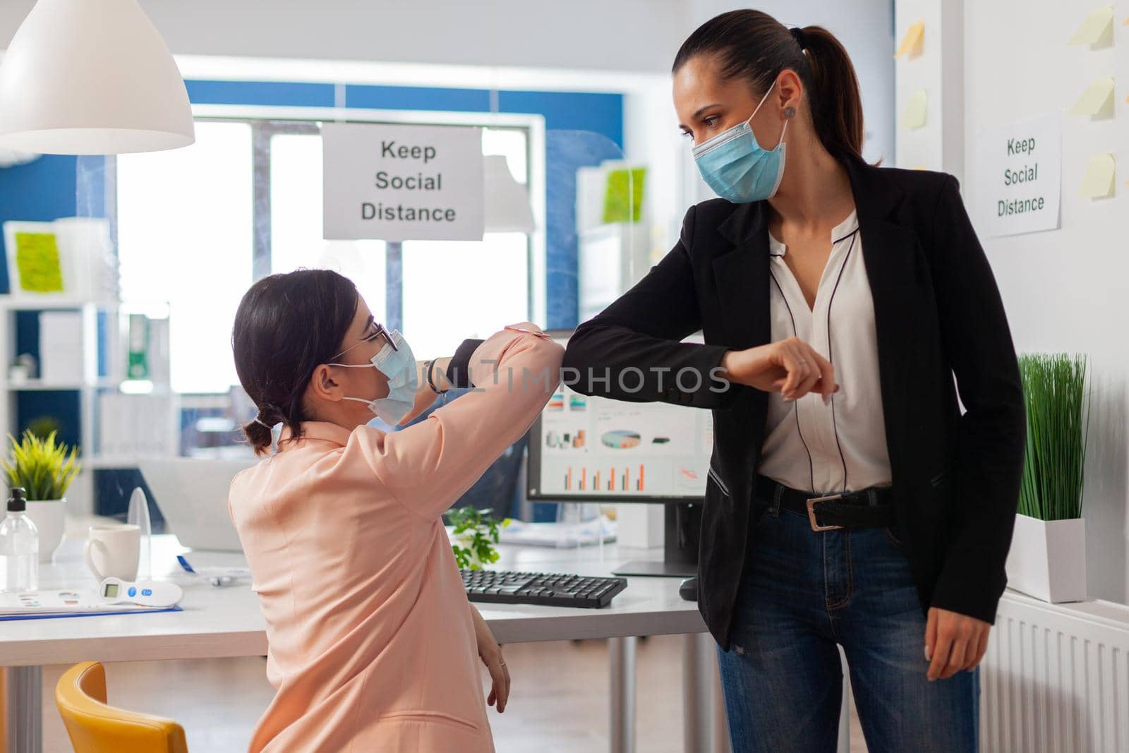 Women greeting at new normal office touching bumping elbows keeping social distancing as safety prevention wearing face mask during global pandemic with covid19.