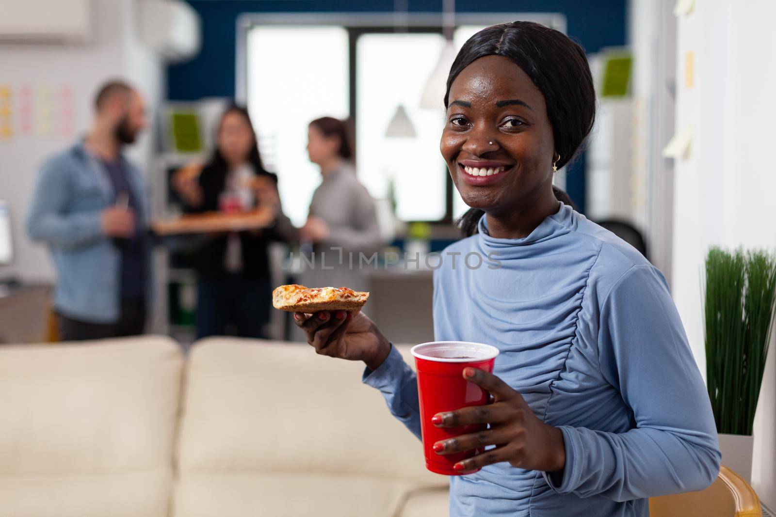 African american woman enjoying food and drinks after work with colleagues. Worker holding slice of pizza and cup with drink alcohol meeting with friends for fun cheerful celebration
