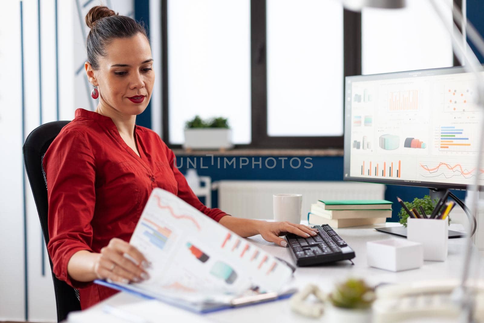 Business woman in start up company working on computer and analyzing charts on clipboard, sitting at desk. Successful concentrated manager working on deadline project typing.