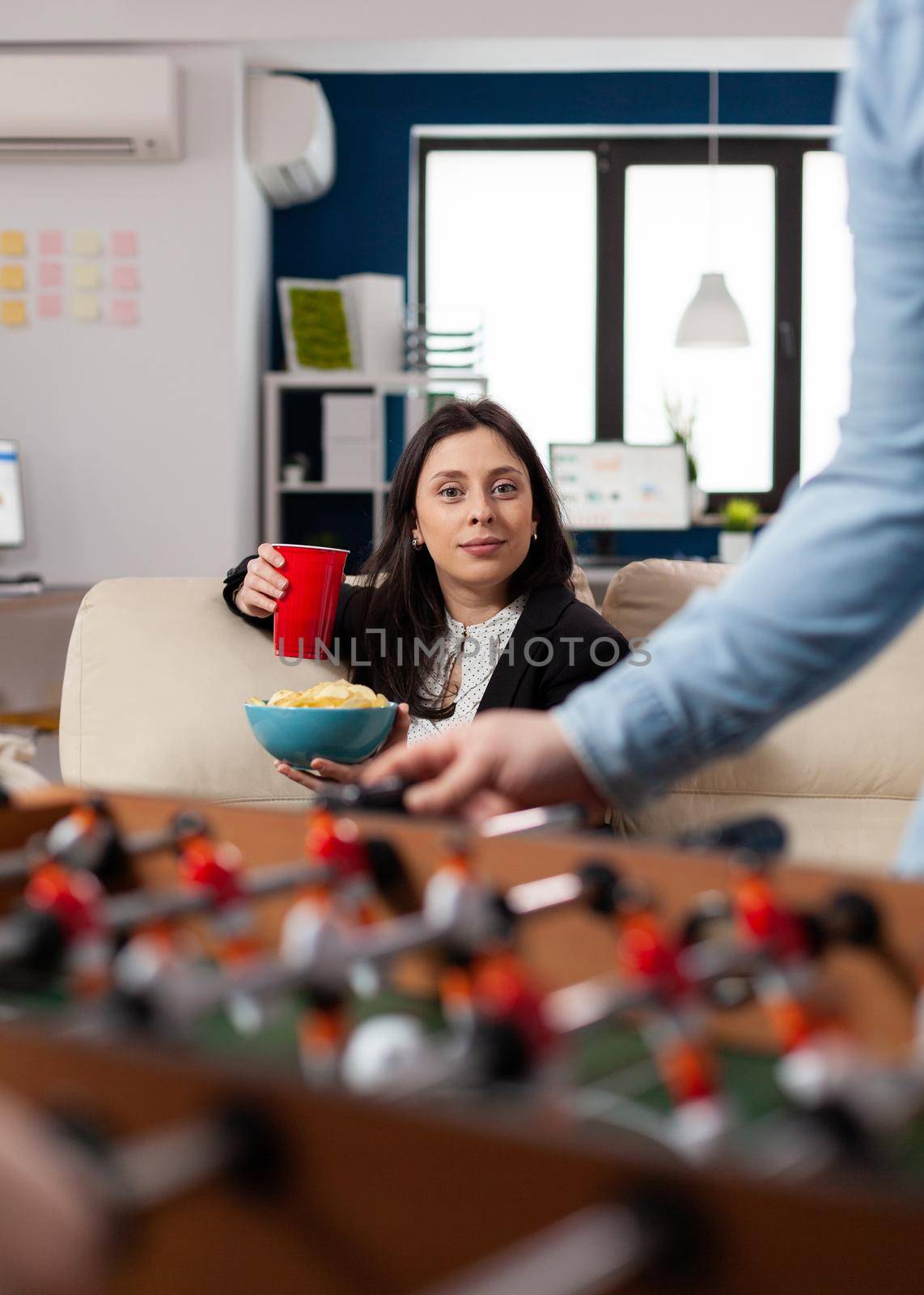 Caucasian woman holding cup of beer and chips after work looking at foosball table soccer game football for fun enjoyment entertainment. Happy worker drinking alcohol at party