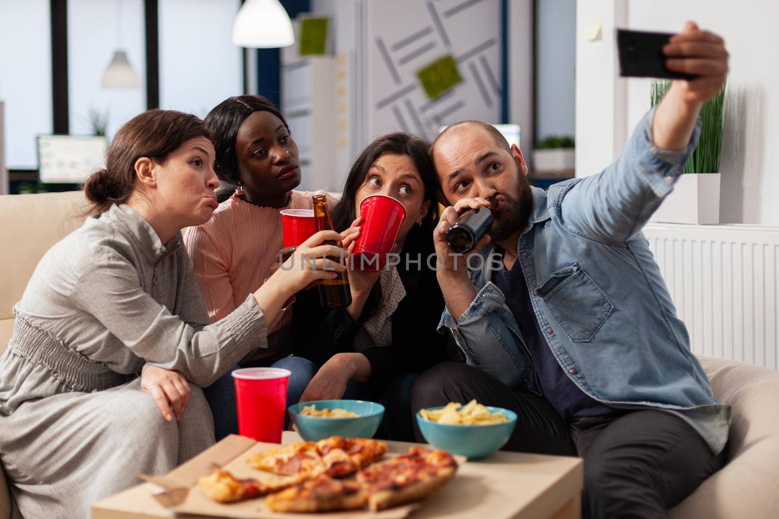 Multi ethnic team of friends make memories at after work party taking pictures on smartphone. Coworkers celebrating at office space with snacks pizza chips cups of drinks on table