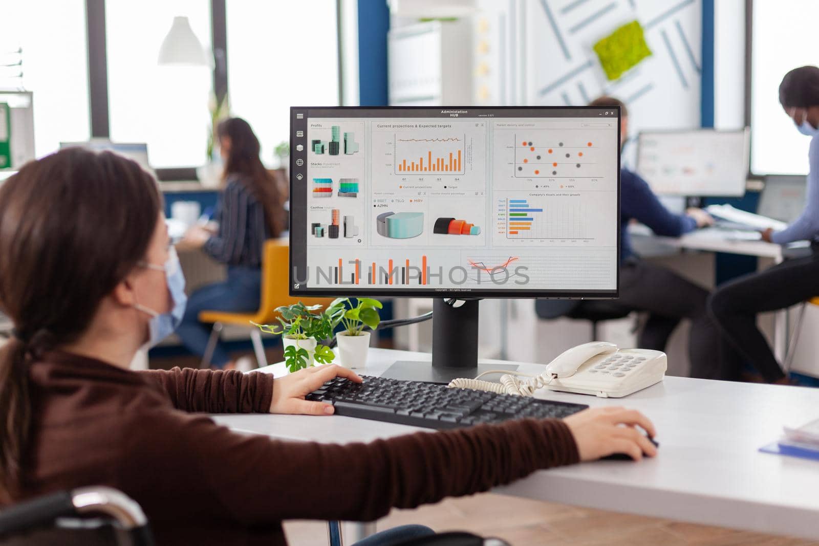 Disabled handicapped businesswoman with face mask immobilized in wheelchair working at financial project typing on pc showing data, prosessing on display sitting in new normal startup business office.