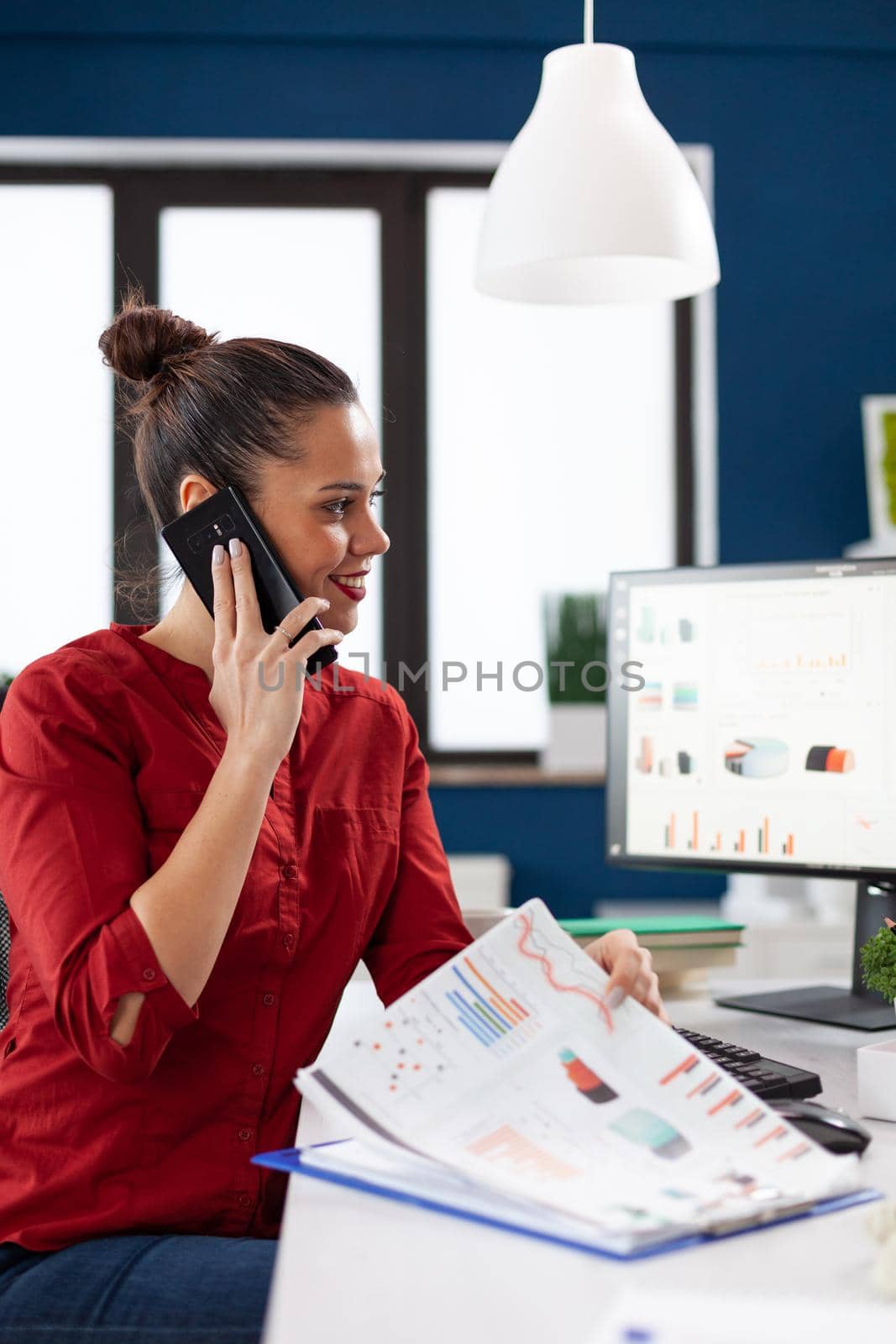 Businesswoman making a call using smartphone sitting at desk in corporate office, looking through financial charts on clipboard. Talking with advisor over phone call giving answers.