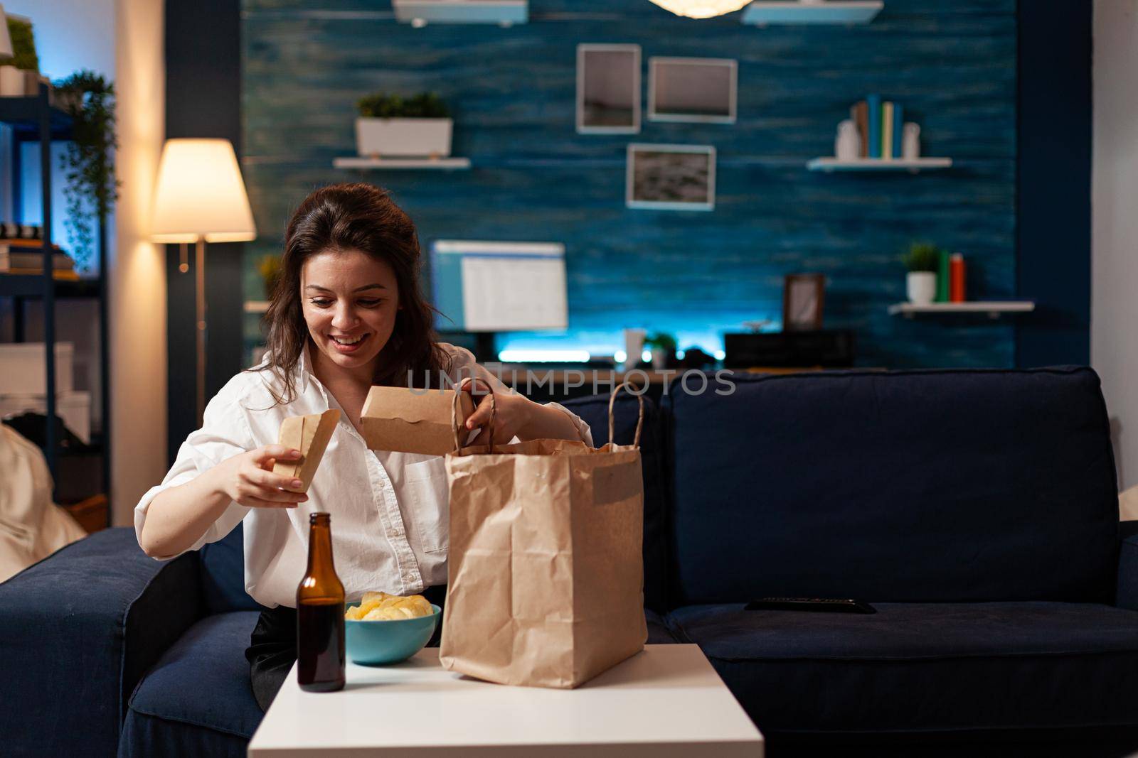 Smiling cheerful woman unpacking tasting fast-food home delivered sitting on couch having lunch serivce order. Caucasian female enjoying delicious takeaway delivery tasty meal in evening