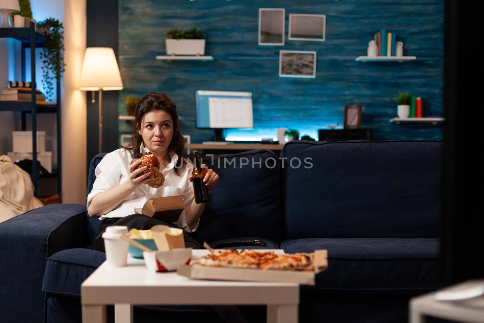 Woman relaxing on couch holding beer bottle while eating tasty delicious burger watching entertainment movie series on television. Caucasian female enjoying takeaway food home delivered at night