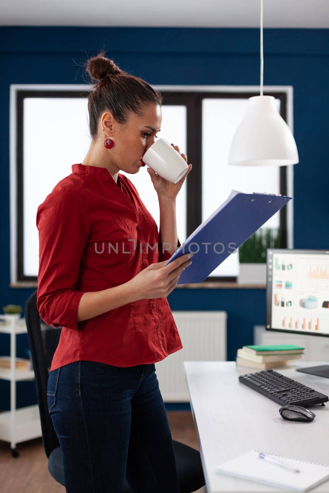 Successful business woman in company office looking through charts while holding and ejoying a cup of tasty coffee. Thoughtfull entrepreneur getting doze of caffeine.