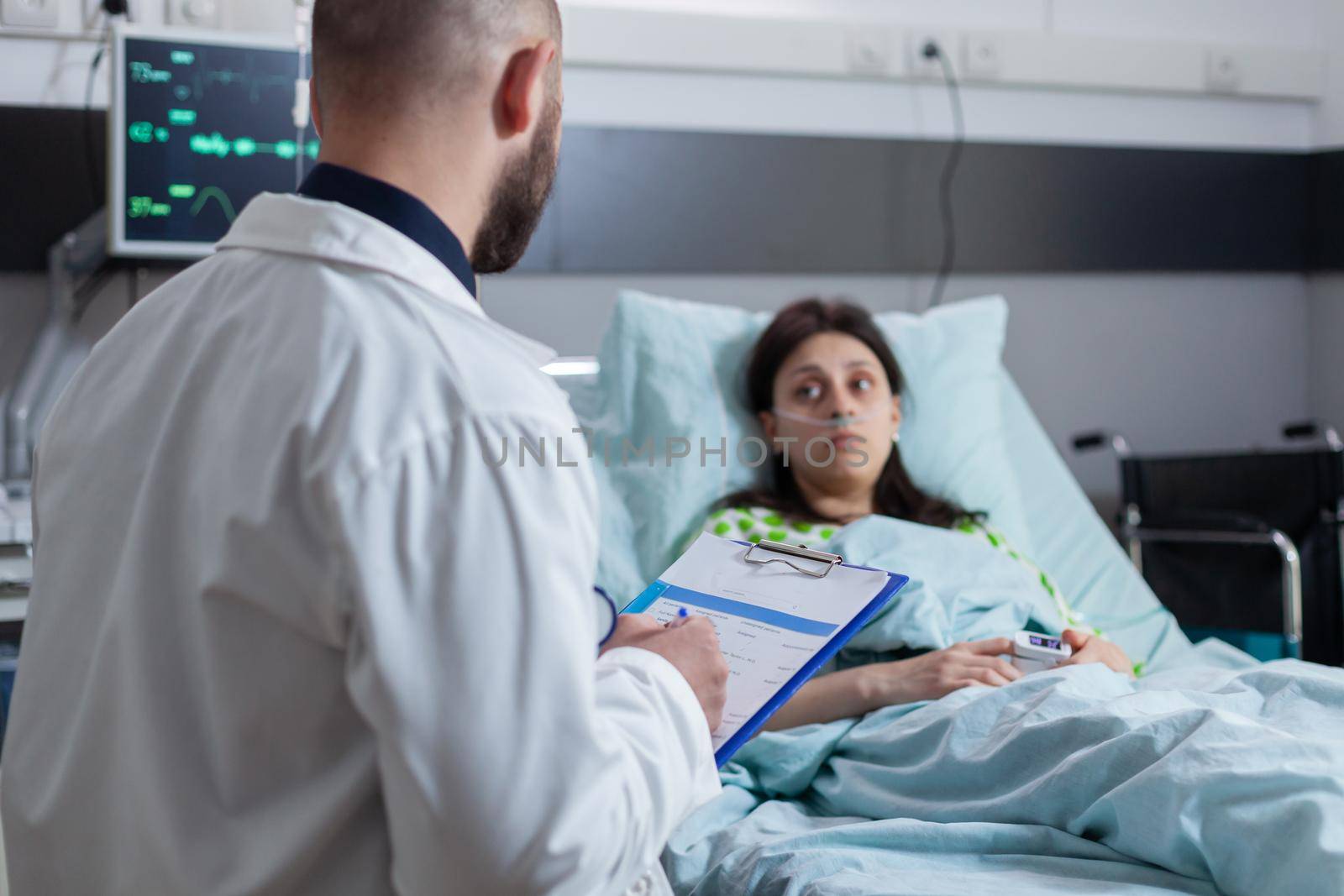 Oncologist practitioner doctor explaining sickness diagnose writing medical disease treatment on clipboard working in hospital ward. Patient sitting in bed recovering after medical surgery, health care system