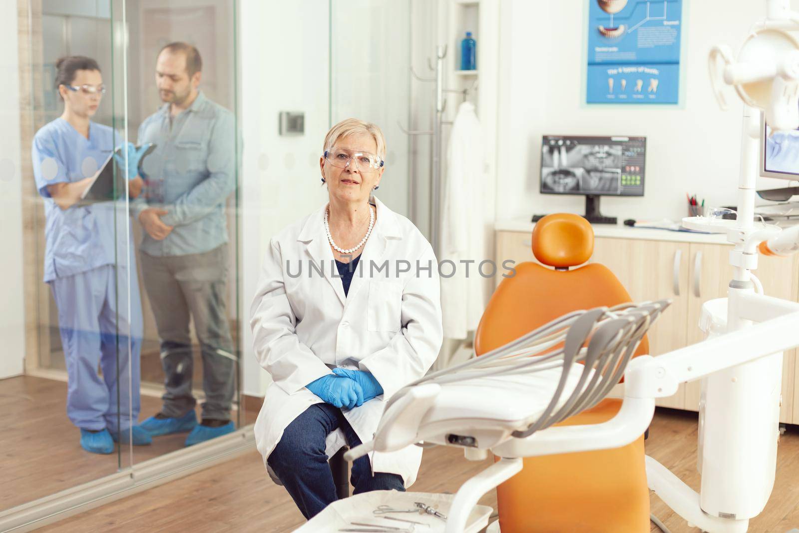 Portrait of smiling senior dentist woman in dental office while medical nurse talking with patient in background. Stomatologist doctor looking on webcam sitting on chair in stomatological clinic