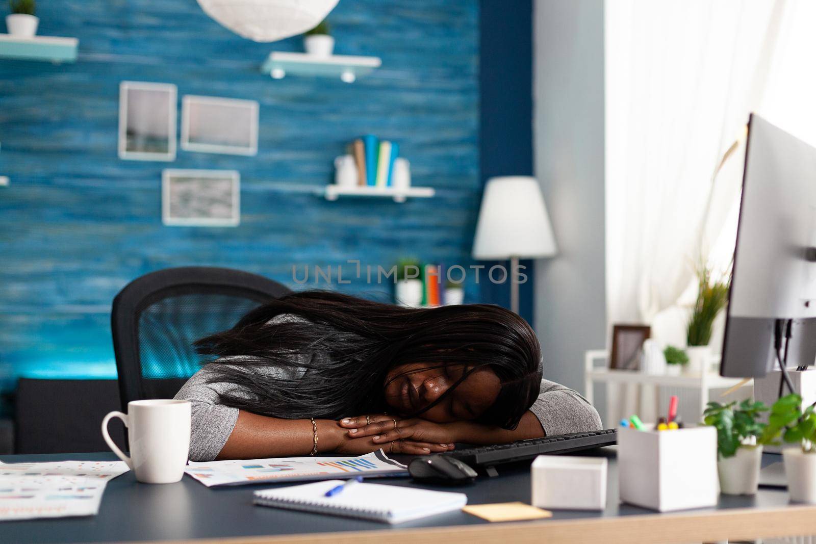 Exhausted tired workaholic black student sleeping on desk table in living room by DCStudio