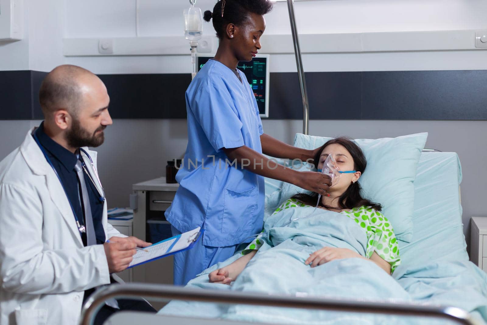 Afro american asisstant putting oxigen mask to sick woman while lying in bed having respiratory disease in hospital ward. Practitioner monitoring heart pulse writing sickness condition on clipboard